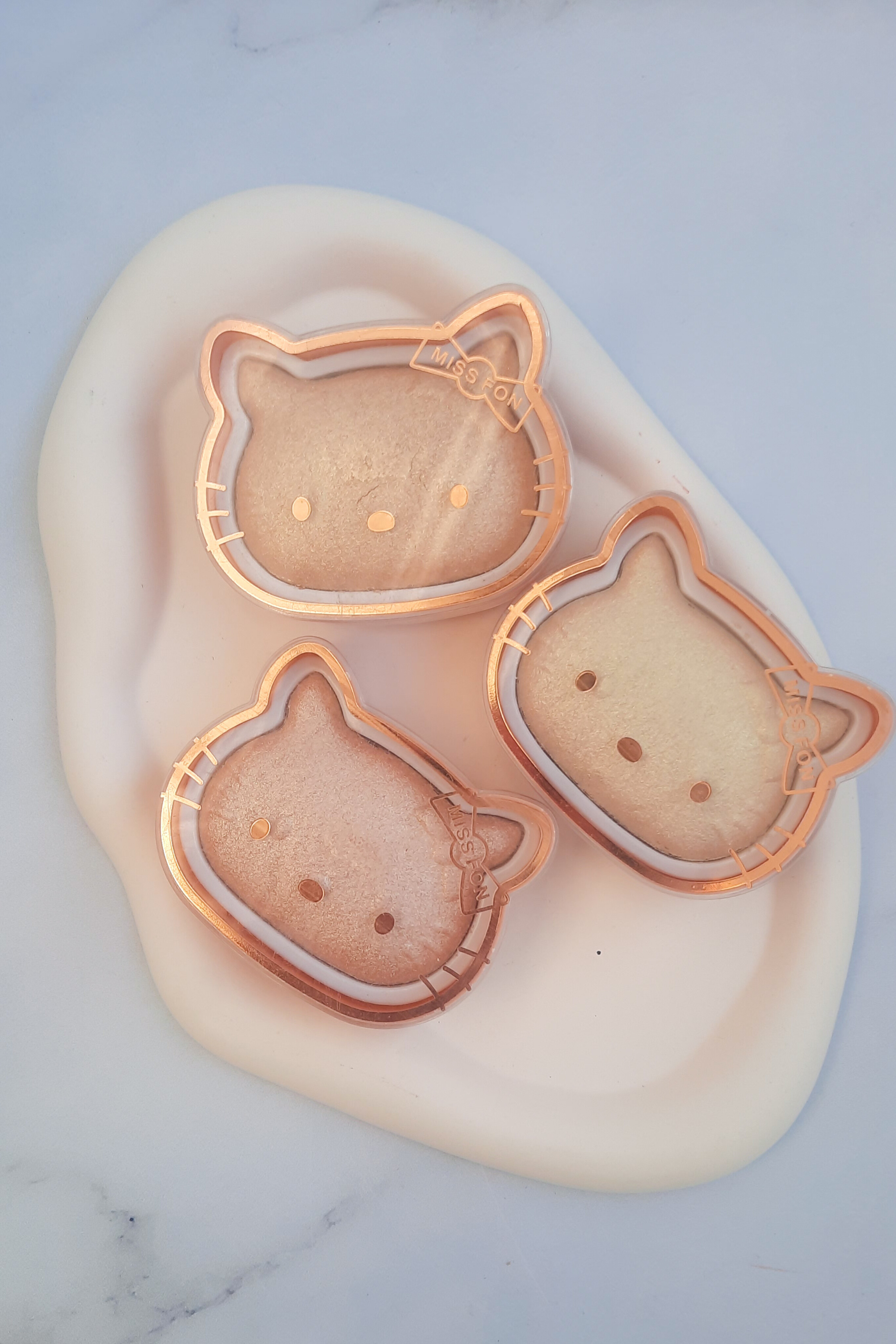 ۳ هایلایتر کیتی MISS Fon کنار هم روی شیپ سنگی سفید دفرمه، رنگ‌های درخشان و طبیعی، بافت نرم و پوشش یکنواخت